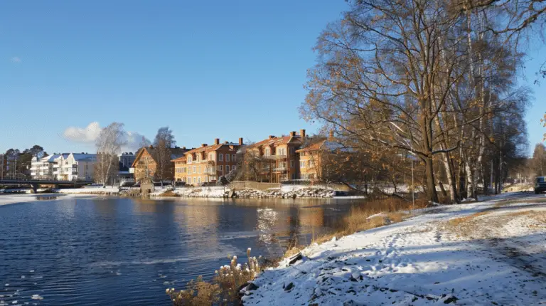 Ett naturskönt vinterlandskap visar en lugn flod kantad av snötäckta stigar och bladlösa träd. På den motsatta stranden ligger en rad pittoreska tegelbyggnader och bostäder, några utrustade med Solceller i Åkersberga, täckta av snö och badade i starkt solljus mot en klarblå himmel.