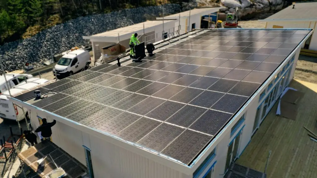 Flygfoto av arbetare i säkerhetsutrustning som installerar solpaneler på det platta taket av en vit byggnad, med sikte på plusenergihus effektivitet. Runt byggnaden finns entreprenadutrustning, en vit skåpbil och grusterräng med en stenig kulle i bakgrunden. Scenen, utspelad i dagsljus, ligger i Norrtälje.