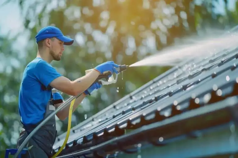 En person som bär en blå keps, blå skjorta och handskar utför en solcellstvätt på ett tak med en högtrycksvattenspray. De står på en stege för säkerhets skull, med solljus som filtrerar genom de omgivande träden.