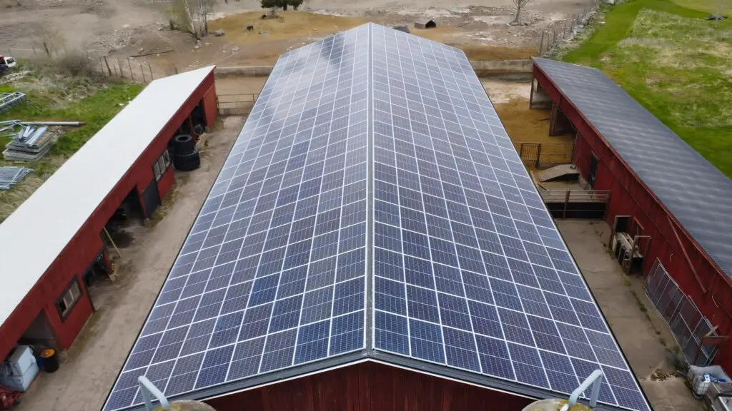 Ett stort ladugårdstak täckt med solceller för lantbruk, omgivet av röda gårdsbyggnader och gräsbevuxna ytor, med fält synliga i bakgrunden.