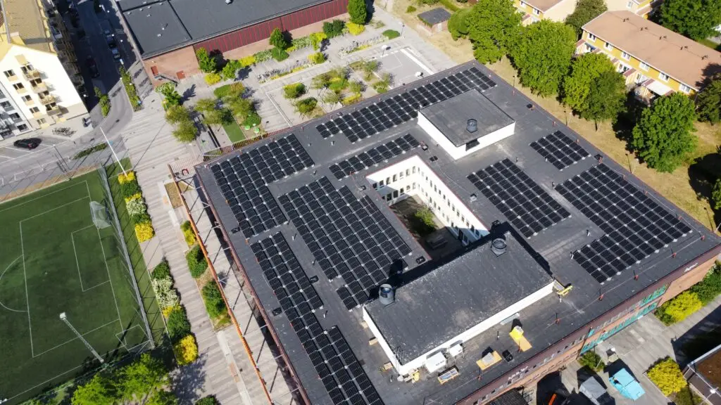 Flygfoto över en stor byggnad med solpaneler och Solcellsbatteri för företag på taket, omgiven av träd, en idrottsplan, parkeringsplats och närliggande byggnader.