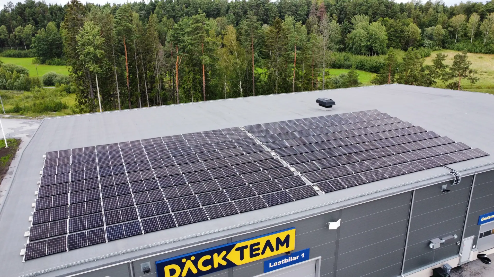 Flygfoto över en byggnad med platt tak, många solpaneler och ett solcellsbatteri för företag installerat ovanpå, omgiven av gröna träd och gräs. Byggnaden har en skylt med texten "Däck Team".