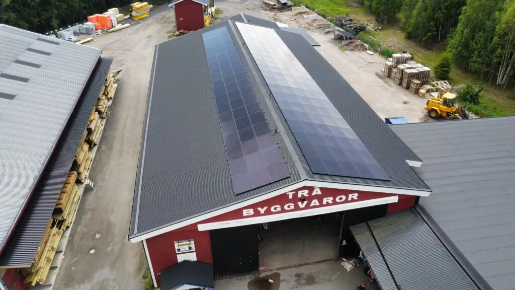 Flygfoto över ett stort rött lager med solpaneler och ett solcellsbatteri för företag på taket, omgivet av industrimaterial, träd och utrustning på en gård.