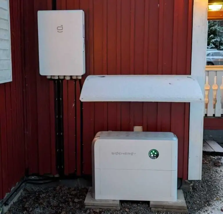 Ett vitt Synergy solcellsbatteri för villa och en monterad elbox är installerade på den röda ytterväggen på en byggnad, delvis täckt av ett litet tak. Taket och marken är lätt snötäckta.